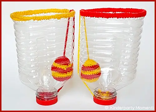 Zwei aufrecht stehende, durchsichtige Plastikflaschen mit gehäkelten roten und gelben Rändern und daran befestigten gehäkelten Bällen in Rot und Gelb