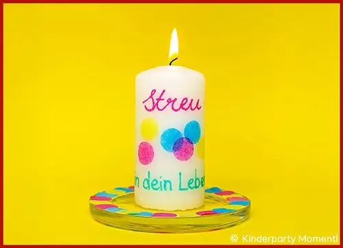 Brennende weiße Kerze mit bunten Punkten und Schrift auf einem mit buntem Konfetti dekorierten Glasuntersetzer vor gelbem Hintergrund
