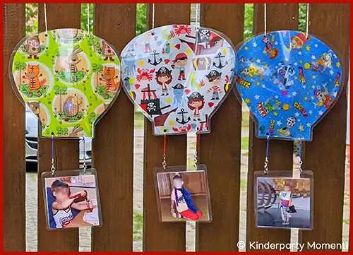 Drei bunte Ballons mit Kinderparty-Motiven hängen an einer Holzwand, jeder Ballon hat ein Foto eines Kindes darunter befestigt