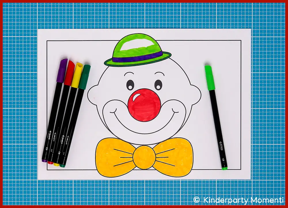Ausgemaltes Clowngesicht mit roter Nase, gelber Fliege und grün-weiß gestreiftem Hut auf weißem Papier, umgeben von Filzstiften auf blauem Schneideunterlage.