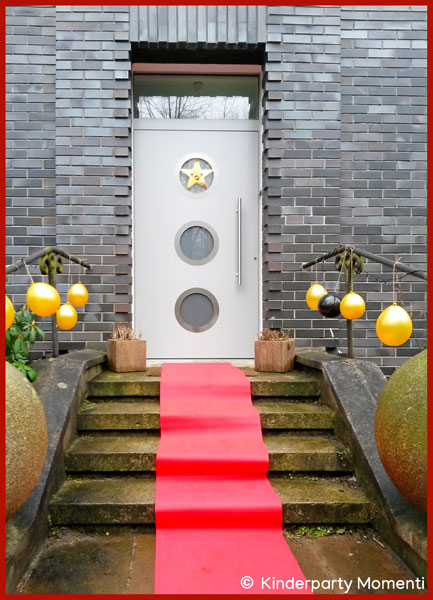Roter Teppich bei Filmgeburtstag auf der Treppe vor einem Hauseingang liegt ein roter Teppich, am Geländer hängen goldene Luftballons