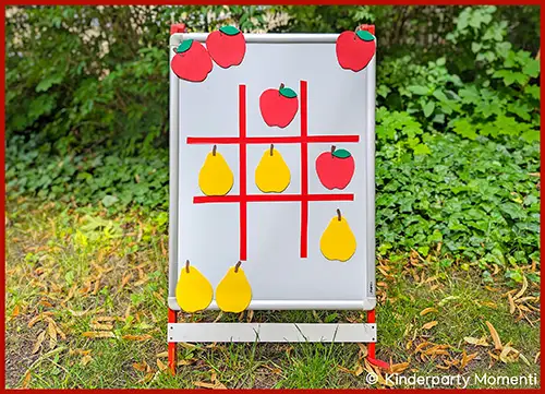 Dreiergitter aus roten Linien auf weißer Tafel mit magnetischen Apfel- und Birnenformen als Spielfiguren im Freien