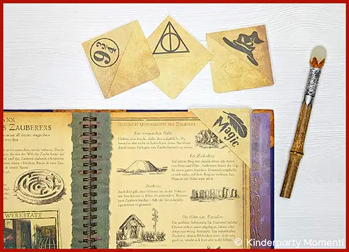 Aufgeschlagenes Zaubererbuch mit Text und Illustrationen, drei goldene Lesezeichen mit Harry-Potter-Symbolen und ein Zauberstab mit silbernem Griffband auf hellem Untergrund