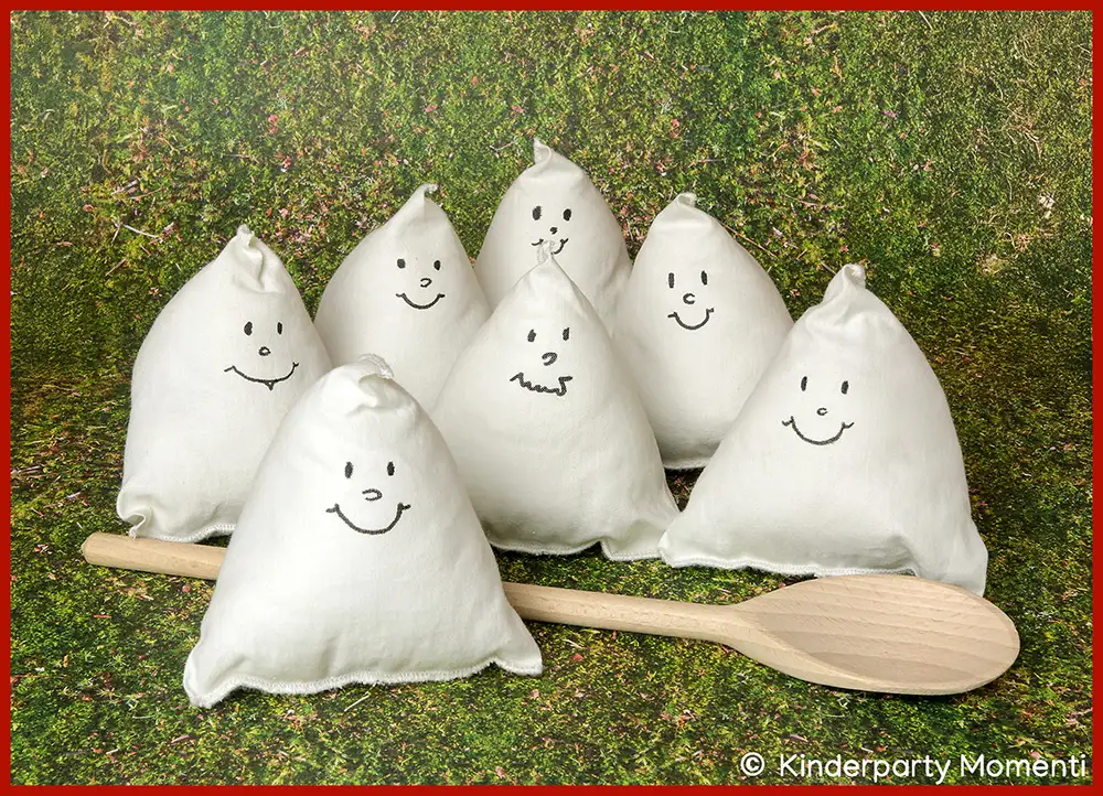 Sieben, kleine, weiße Stoff Gespenster in Dreiecksform mit aufgemalten, lachenden Gesichtern und ein großer Holzlöffel auf grünem Hintergrund.