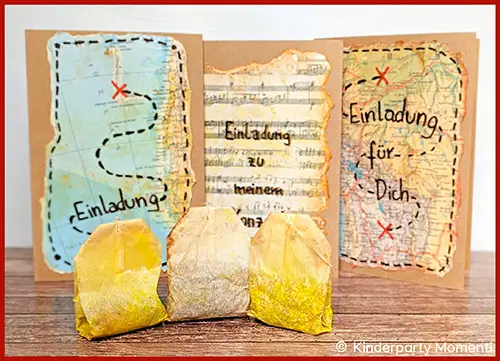 Drei handgemachte Einladungskarten mit Schatzsuche-Motiven und handgeschriebener Schrift auf einer Holzoberfläche, davor drei gelb gefärbte Teebeutel