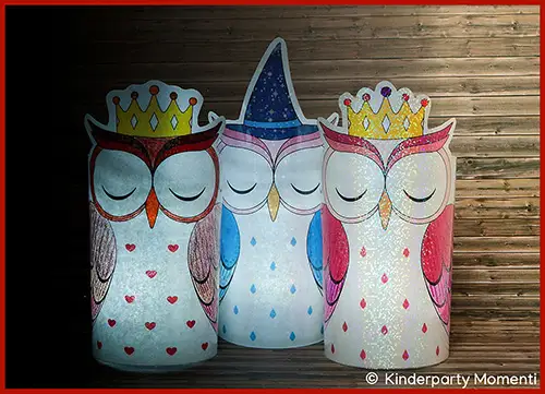 Drei leuchtende Eulen DIY Windlichter aus Papier, zwei mit goldener Krone und eine mit lila Hexenhut, vor Holzwand.