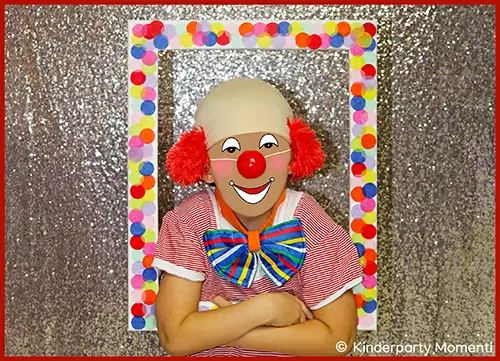 Kind mit rotem Clownshaar, rotem gepunktetem Hemd und großem mehrfarbigem Fliegenknoten vor einem rechteckigen Bilderrahmen mit buntem Konfetti auf glitzerndem Hintergrund