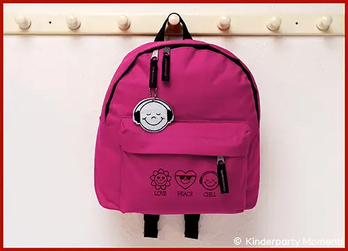 Pinkfarbener Rucksack mit drei aufgedruckten Symbolen und den Worten 'Love Peace Chill' sowie einem Anhänger in Form eines lächelnden Gesichts mit Kopfhörern