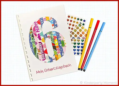 Gebundenes Buch mit einer großen Zahl 6 aus bunten Geschenkpapierstreifen auf dem Cover und der Aufschrift 'Mein Geburtstagsbuch' daneben bunte Stifte und Aufkleberbögen mit Sternen, Blumen und Herzen