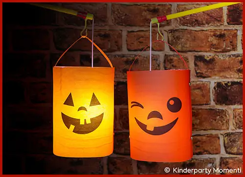 Zwei orangefarbene Halloween-Laternen aus Papier mit lachenden Gesichtern hängen vor einer Ziegelsteinwand.