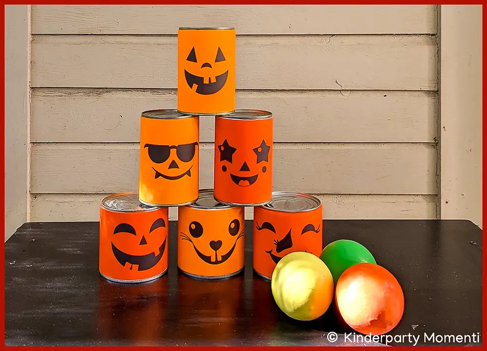 Dosenwerfen Spiel aus sechs Dosen mit orangefarbenen Halloween-Kürbisgesichtern auf schwarzem Tisch, daneben drei bunte Bälle
