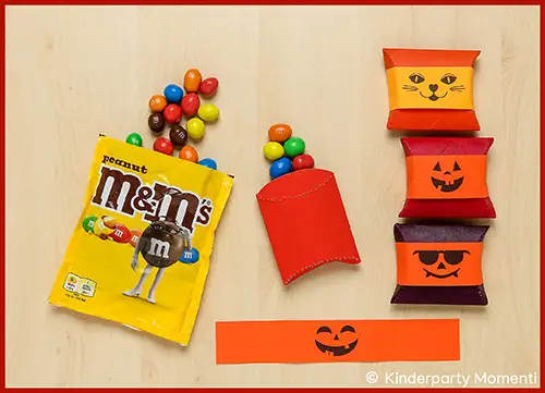 Geöffnete gelbe M&M'S-Tüte mit bunten Schokolinsen daneben und vier kleine Kissenschachteln aus Klopapierrollen mit Halloween-Motiven in Orange, Rot und Lila