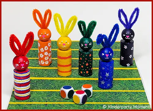 Sechs bunte Hasenfiguren aus Holz mit Ohren aus Pfeifenreinigern stehen auf einem Spielfeld mit grüner Oberfläche und gelben Linien, davor liegen drei Würfel mit bunten Punkten