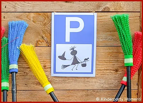 Blaues Parkplatzschild mit weißem P und schwarzer Hexenfigur auf Besen, umgeben von bunten Besen vor Holzwand
