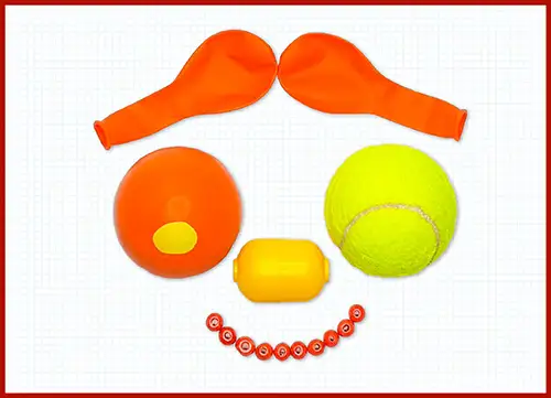 Aus verschiedenen orangenen und gelben Gegenständen gelegtes Gesicht mit zwei Luftballons als Augen und einer Tennisball als Nase