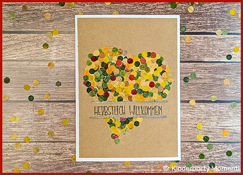 Herz aus buntem Konfetti auf braunem Papier mit Schriftzug 'HERBSTLICH WILLKOMMEN' auf Holztisch mit verstreutem Konfetti