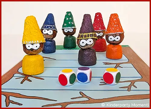 Sechs Kastanienfiguren mit bunten spitzen Hüten und großen weißen Augen stehen auf einem Spielfeld mit Astmotiven, davor liegen drei bunte Würfel
