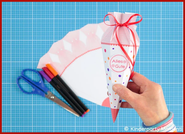 Eine Hand hält eine kleine selbstgebastelte Schultüte auf der "Alles Gute" steht. Im Hintergrund ist die Schultüten Vorlage und Bastelwerkzeug zu sehen.