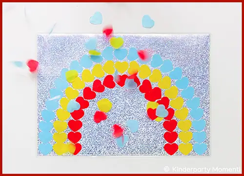 Rechteckiges, glitzerndes Tischset mit einem Regenbogen aus roten, gelben und blauen Herzkonfetti