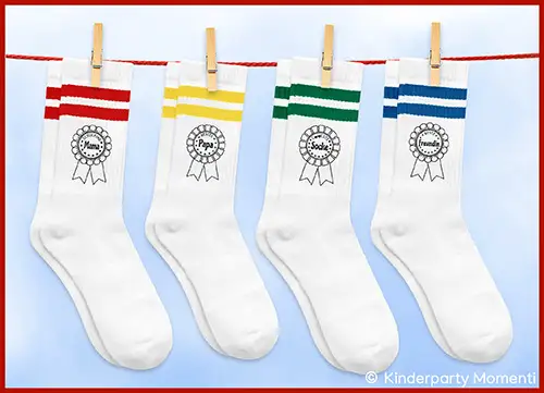 Vier weiße Socken mit farbigen Streifen oben und Aufdrucken 'Schulkind Mama', 'Bester Papa', 'Coolste Socke', 'Beste Freundin' hängen an einer Wäscheleine mit Wäscheklammern
