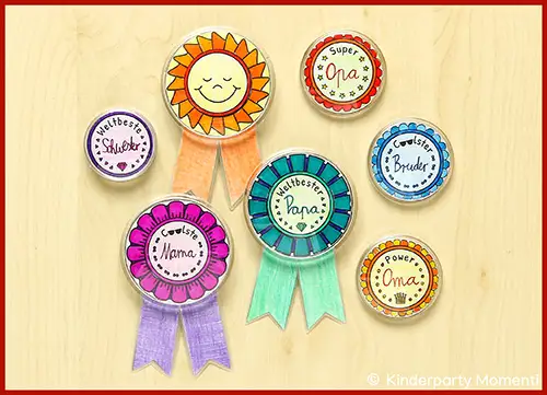 Sieben bunte, handgezeichnete Orden mit Sonnen- und Blumenmotiven und Texten wie 'Weltbeste Schwester', 'Coolster Bruder', 'Weltbester Papa', 'Coolste Mama', 'Super Oma' und 'Power Oma' auf hellem Holzuntergrund