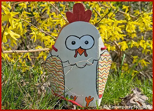 Aus Milchkarton gebasteltes Huhn mit rotem Kamm und gemusterten Flügeln steht auf Gras vor gelb blühendem Strauch