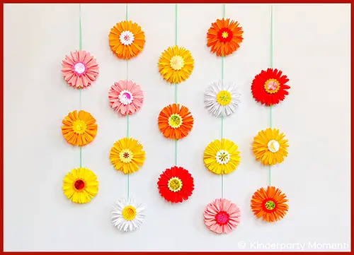 Mehrere bunte Papierblumen in einer Girlande an grünen Fäden hängend vor weißem Hintergrund