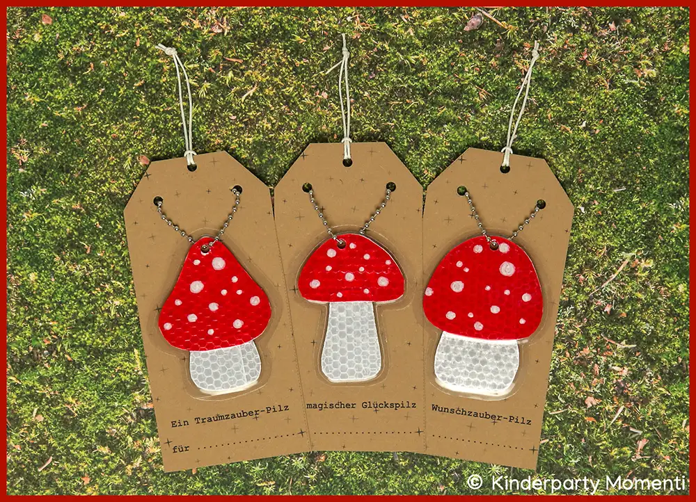 Drei gebastelte Fliegenpilz Anhänger mit roten Kappen und weißen Punkten auf braunen Geschenkanhängern, liegend auf grünem Moos.