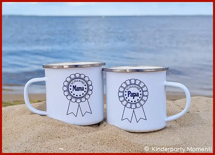 Zwei weiße Emaille-Tassen mit Metallrand im Sand am Wasser, bedruckt mit schwarz-weißen Rosetten und den Schriftzügen 'Schulkind Mama' und 'Schulkind Papa'