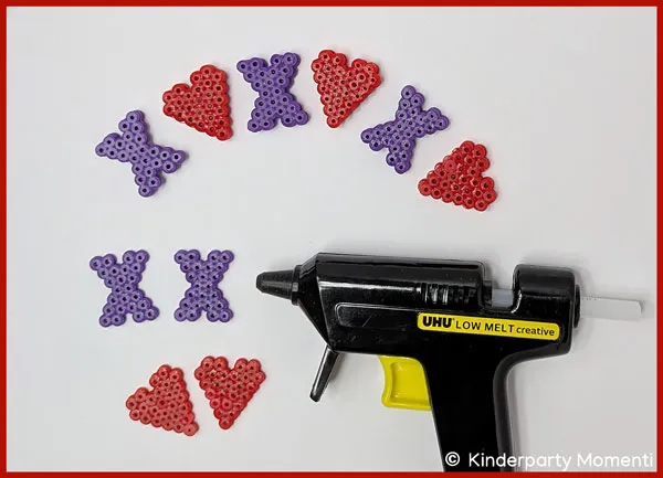 DIY Spielfiguren aus Bügelperlen auf einem Tisch liegen Herzen und Kreuze aus Bügelperlen sowie eine Heißklebepistole