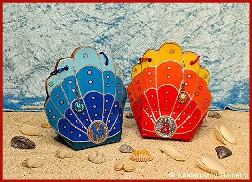Zwei muschelförmige Tetrapak-Tüten in Blau und Rot mit Verzierungen stehen auf Sand umgeben von kleinen Muscheln