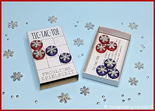 Tic-Tac-Toe-Spiel mit roten und blauen Spielsteinen mit weißen Schneeflockenmotiven auf hellblauem Untergrund, Spielbrett und Schachtel mit der Aufschrift 'TIC-TAC-TOE' und 'FRÖHLICHE SPIELEZEIT'