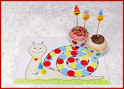 Kindertisch mit Schnecken-Spielbrett aus bunten Punkten, zwei Cupcakes mit Smiley-Picker und Spielfiguren