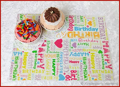 Tischset aus Geschenkpapier mit buntem Happy-Birthday-Schriftzug und zwei Muffins darauf