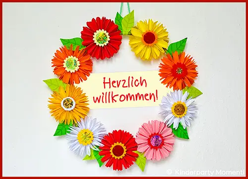 Bunter Papierblumenkranz mit grünen Blättern um ein Schild mit roter Schrift 'Herzlich willkommen!'