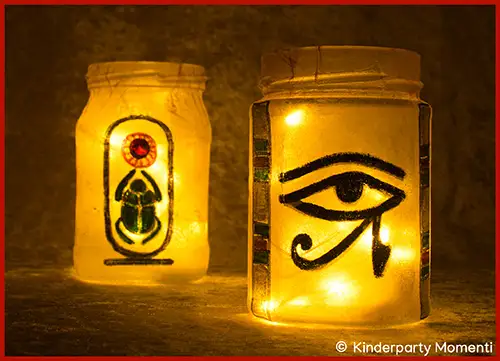 Zwei Windlichtgläser mit leuchtenden Kerzen, eines mit einem ägyptischen Hieroglyphen-Auge, das andere mit einem Skarabäus-Symbol