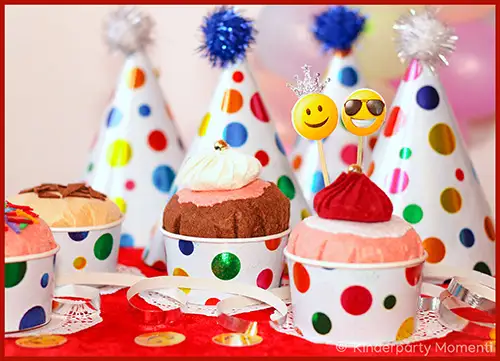 Mehrere bunte Muffins mit Deko und Partyhütchen mit Punkten und Bommel im Hintergrund auf rotem Tuch