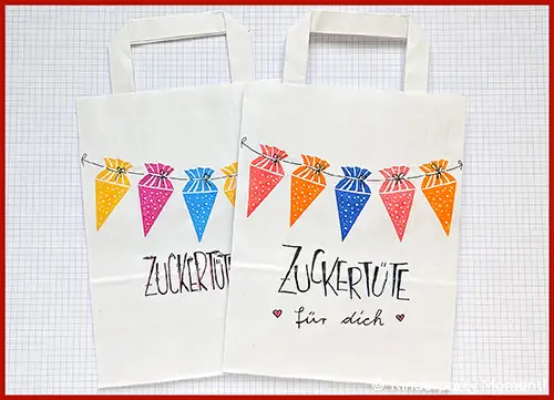 Zwei weiße Papiertüten mit bunten Zuckertüten-Motiven und der Aufschrift 'ZUCKERTÜTE' auf kariertem Hintergrund