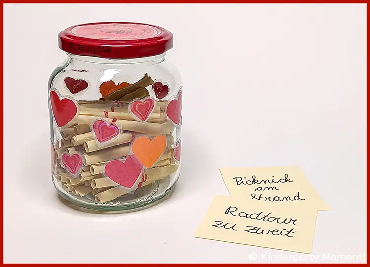 Gutschein Glas mit DIY Herz Sticker Deko Glas mit roten und orangenen Herzstickern beklebt, gefüllt mit zusammengerollten Gutschein-Zetteln , daneben zwei handbeschriebene gelbe Zettel