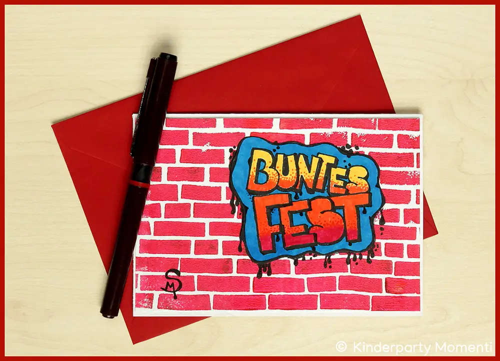 DIY Weihnachtskarte mit rotem Ziegelmauer-Hintergrund und bunter Graffiti-Schrift 'BUNTES FEST', daneben ein schwarzer Stift und ein roter Umschlag.