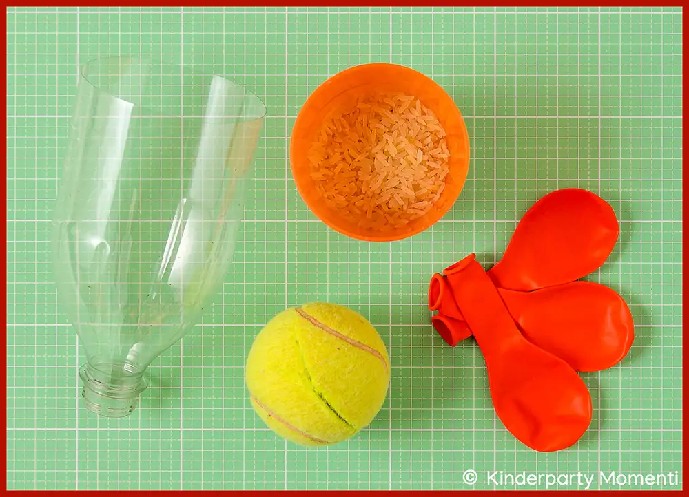 Bastelmaterial für Dosenwerfen Bälle - Zerschnittene Plastikflasche, orangefarbener Becher mit Reis, gelber Tennisball und drei rote Luftballons auf grün kariertem Untergrund