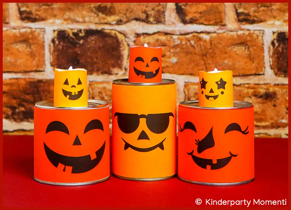 Drei orangefarbene Dosen mit schwarzen Halloween-Kürbisgesichtern, darauf jeweils eine brennende LED Teelichter mit passenden Gesichtern, vor rotem Untergrund und Backsteinwand.