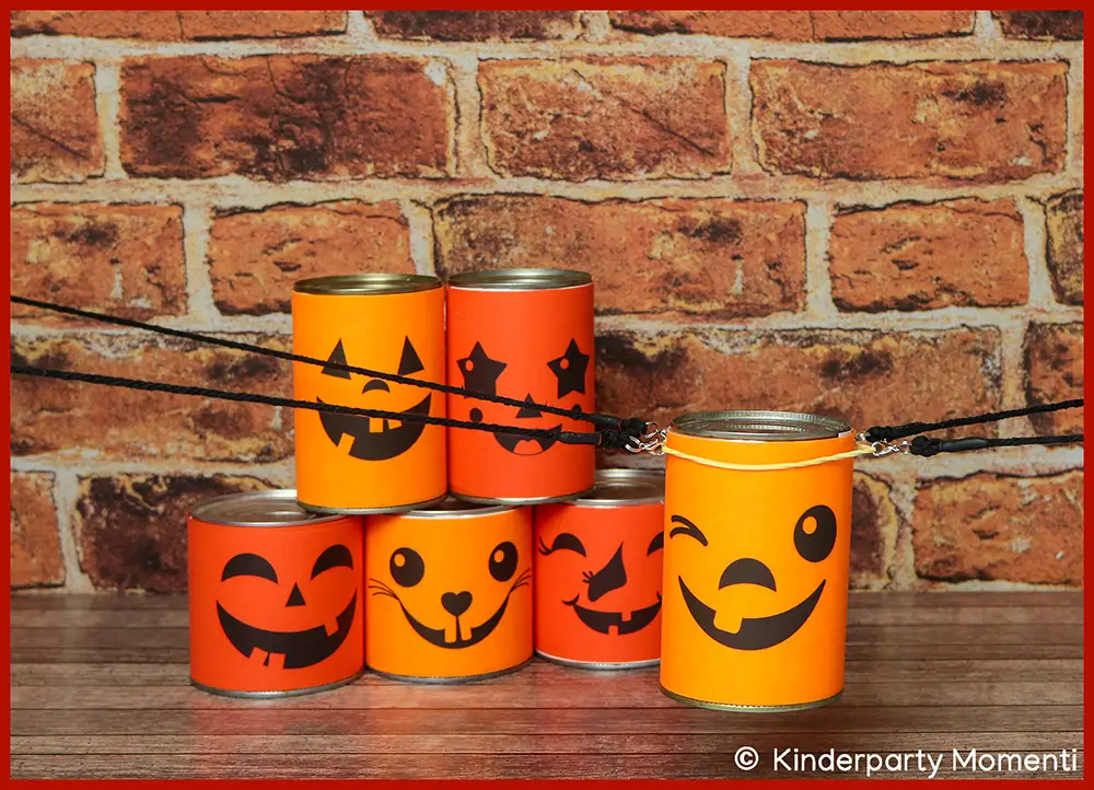 Idee für Kindergeburtstag DIY Spiele für Gruppen - gemeinsam Pyramide aus Bechern stapeln Fünf Dosen mit Neon Papier mit Halloween-Gesichtern beklebt vor roter Backsteinwand, auf Holztisch gestapelt. Eine gleiche Dose schwebt an vier Bändern im Vordergrund