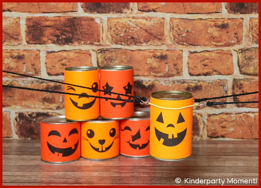 Becher Stapeln Spiel mit Dosen basteln für Gruppenspiele beim Kindergeburtstag Fünf Dosen mit Neon Papier mit Halloween-Gesichtern beklebt vor roter Backsteinwand, auf Holztisch gestapelt. Eine gleiche Dose schwebt an vier Bändern im Vordergrund
