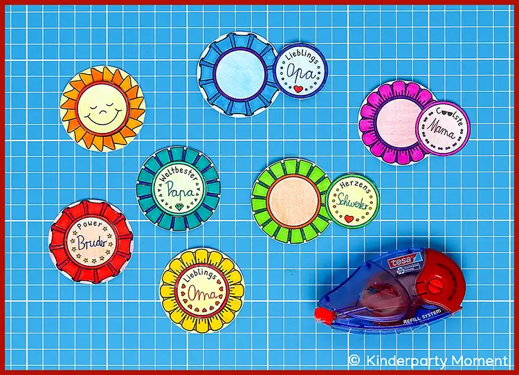Bunte, handgemachte Button mit verschiedenen Beschriftungen wie 'Lieblings Oma', 'Coolste Mama', 'Weltbester Papa', 'Herzens Schwester' und 'Power Bruder' auf kariertem Hintergrund neben einem Kleberoller