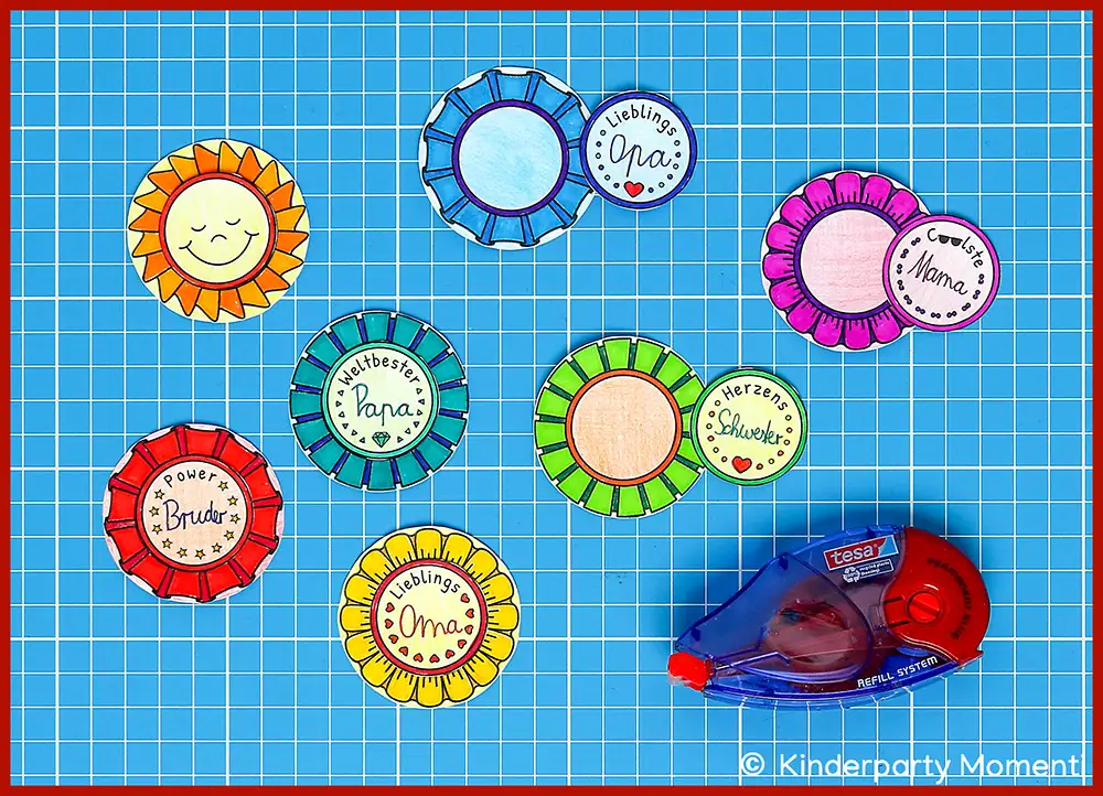 Bunte, handgemachte Button mit verschiedenen Beschriftungen wie 'Lieblings Oma', 'Coolste Mama', 'Weltbester Papa', 'Herzens Schwester' und 'Power Bruder' auf kariertem Hintergrund neben einem Kleberoller