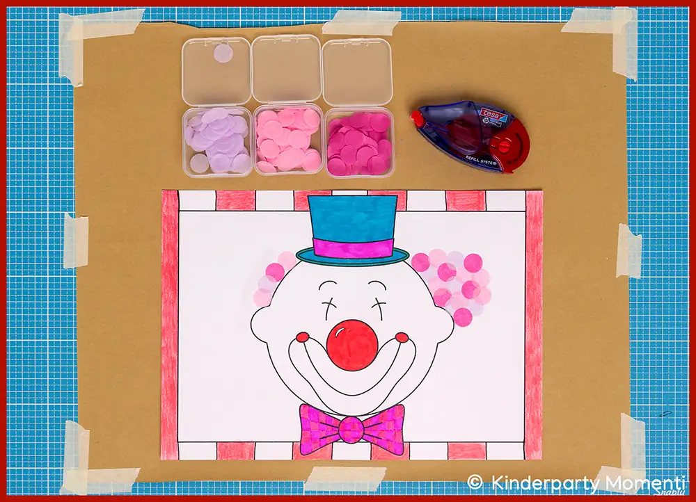 Ausmalbild eines Clownsgesichts mit roter Nase, lila Fliege und blauem Zylinder auf weißem Papier, umgeben von kleinen Behältern mit bunten Konfetti und einem Kleberoller auf einem blauen Schneidunterlage.
