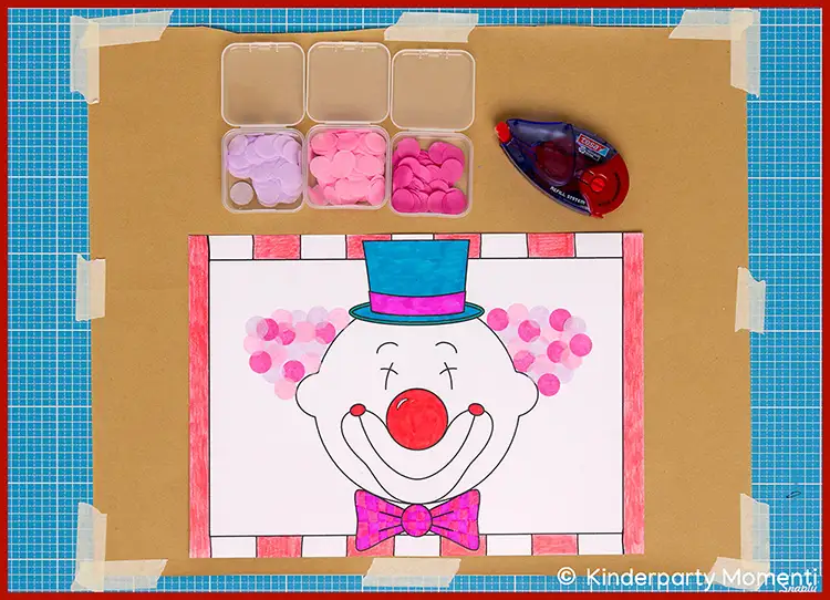Ausmalbild eines Clownsgesichts mit roter Nase, lila Fliege und blauem Zylinder auf weißem Papier, umgeben von kleinen Behältern mit bunten Konfetti und einem Kleberoller auf einem blauen Schneidunterlage.