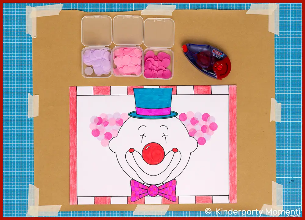 Ausmalbild eines Clownsgesichts mit roter Nase, lila Fliege und blauem Zylinder auf weißem Papier, umgeben von kleinen Behältern mit bunten Konfetti und einem Kleberoller auf einem blauen Schneidunterlage.