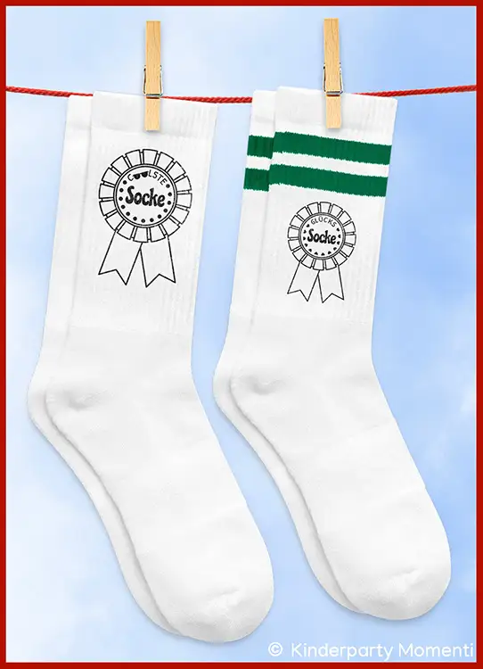 Zwei weiße Socken, eine mit grünem Streifen und Aufdruck einer Siegerrosette mit dem Text 'Coolste Socke' und 'Glückssocke' hängen an einer Wäscheleine